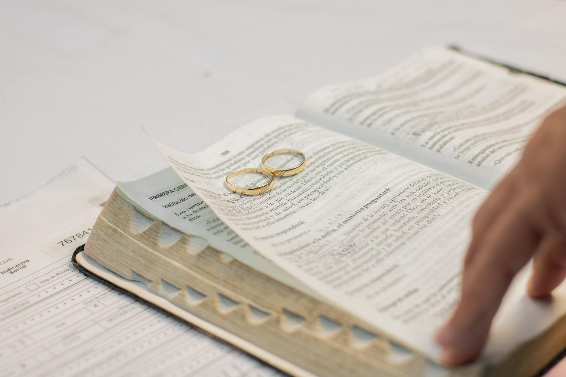 Gold wedding rings rest on an open Bible with a hand turning the page, symbolizing sacred vows.