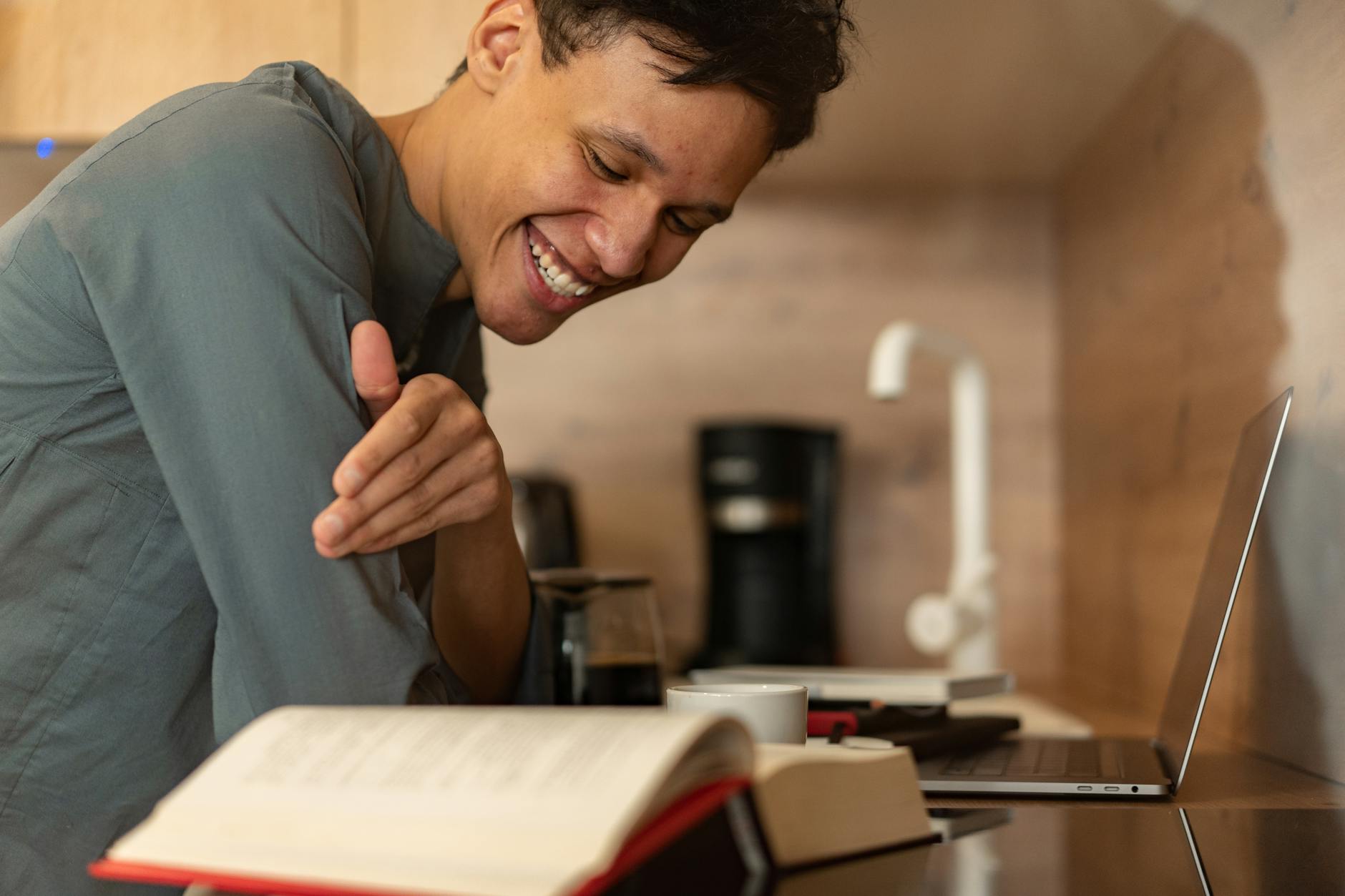 Cheerful man smiling while reading a book with a laptop and coffee indoors, creating a cozy atmosphere.