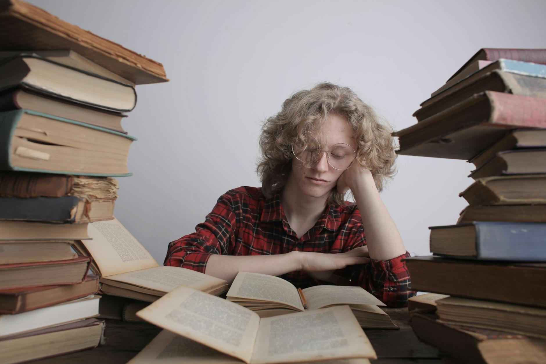 Weary male teenager with long blond hair in casual outfit and eyeglasses sitting at table in library with pile of various books propping up head with hand and reading book during daytime
