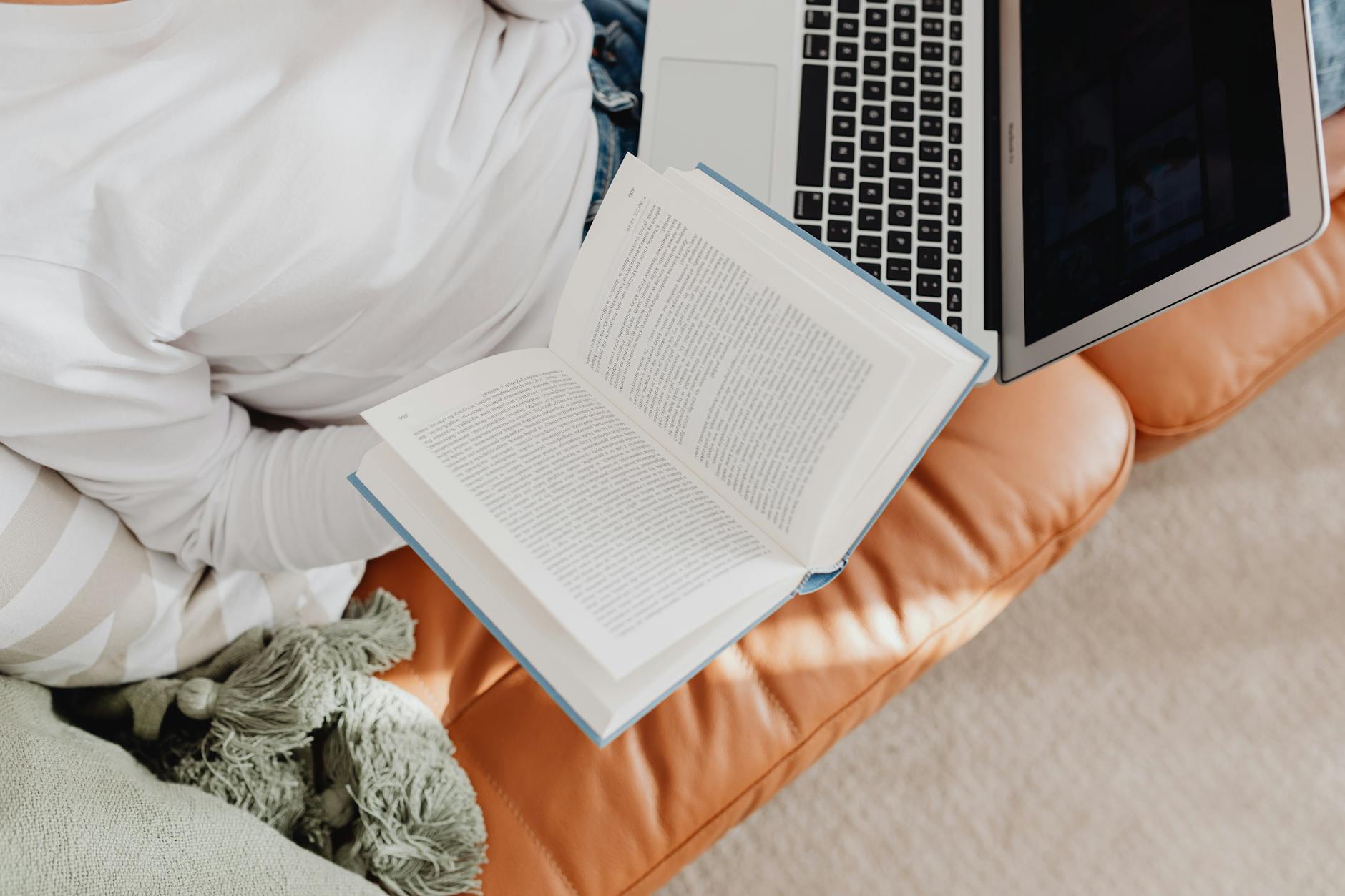 Person on couch reading a book with a laptop nearby, showcasing relaxation at home.